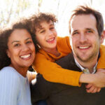 Mum, dad and young boy. Boy is hugging dad round the neck. All are smiling at the camera